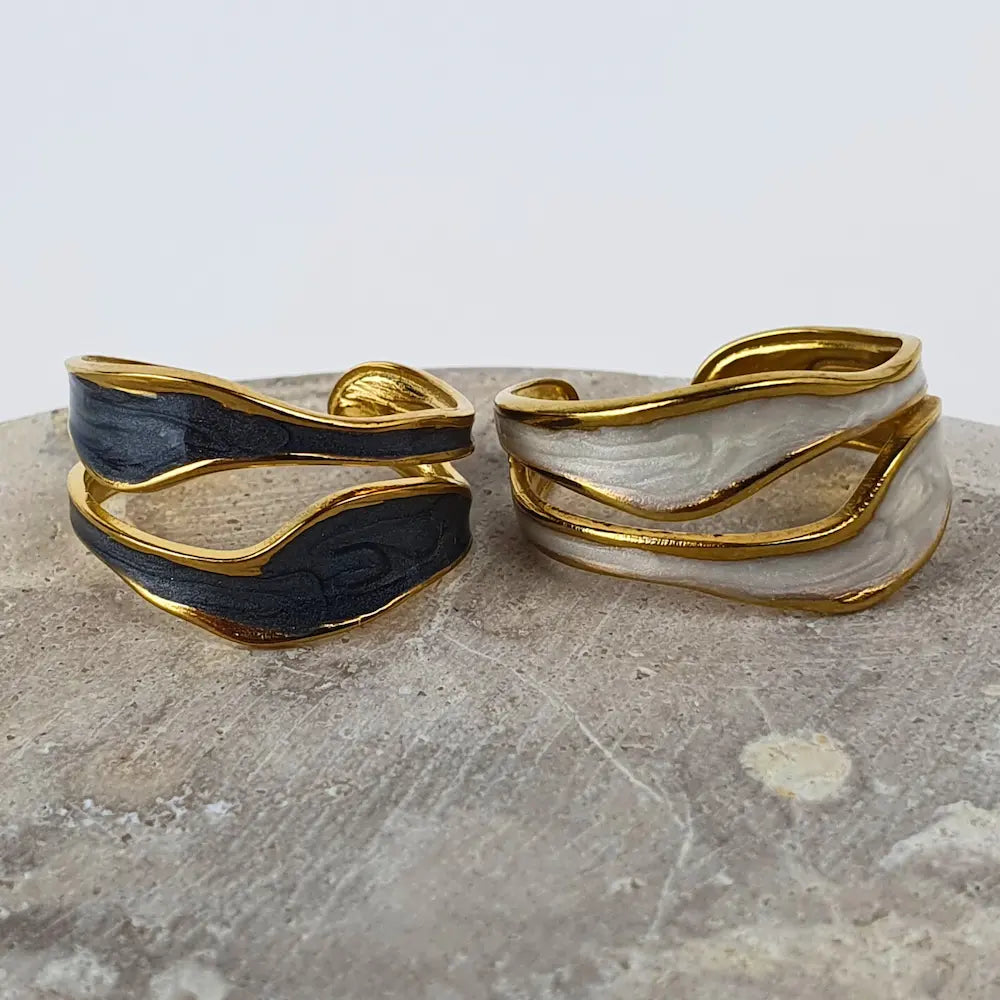 Two Flowing Waters Adjustable Rings shown in 18K gold-plating, one with black oil-dripping and one with white, designed to fit any finger and add bold elegance to any look.