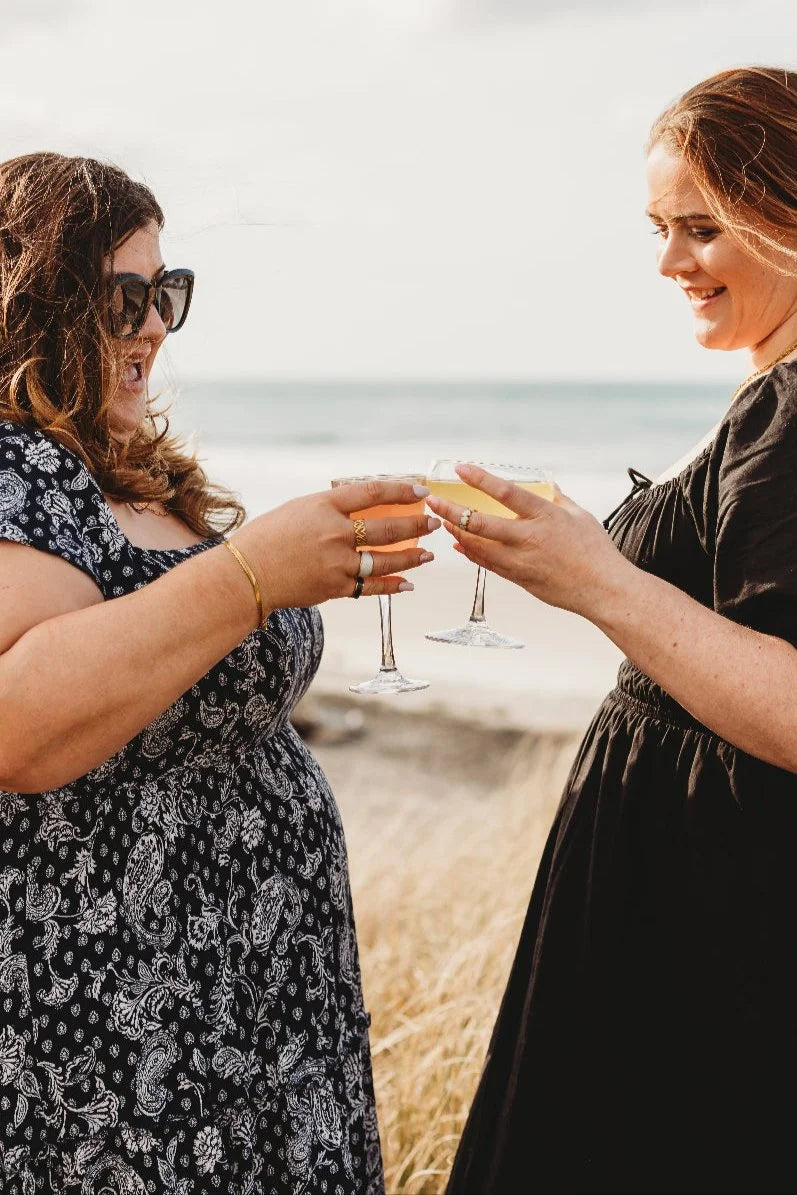 Two women celebrating with drinks on a beach, wearing size-inclusive, adjustable jewellery designed for every body.