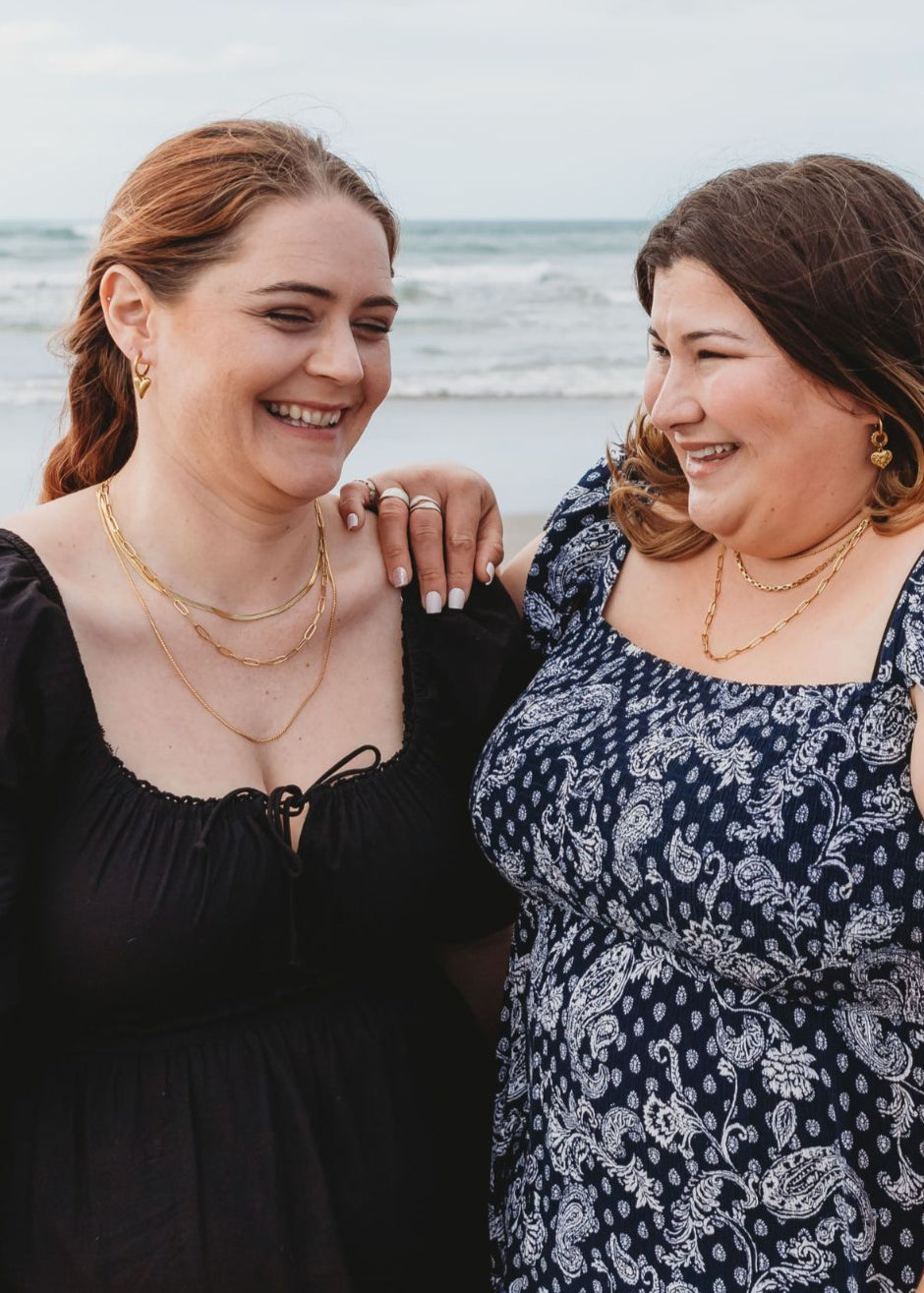 Two diverse friends laughing together on a sunny NZ beach, proudly wearing Divine Me jewellery. Designed for every body - stylish, empowering, and made to fit.