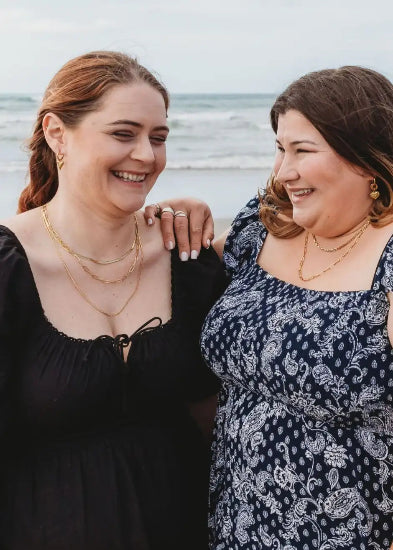 Two diverse friends laughing together on a sunny NZ beach, proudly wearing Divine Me jewellery. Designed for every body - stylish, empowering, and made to fit.